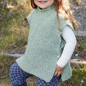 Op de afbeelding: Een lichtgroene gebreide poncho met een hoge kraag en knoopsluiting. De mouwloze poncho heeft een textuur gebreid patroon. Het kind draagt een wit shirt met lange mouwen en een blauwe broek met patroon. De achtergrond is wazig.