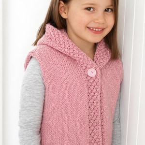 May include: A pink knitted hooded vest with a large button closure. The vest has a textured border and a matching hood. The child is wearing a gray long-sleeved shirt underneath the vest.