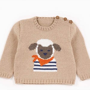 Op de afbeelding: Beige gebreide trui met een schapenmotief. Het schaap heeft een wit gezicht, bruine oren en draagt een blauw-wit gestreept shirt met een oranje sjaal. De trui heeft drie houten knopen op de schouder.