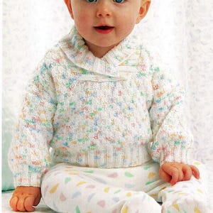 May include: A baby wearing a white knit sweater with a cowl neck and pastel-colored flecks. The baby is also wearing white footed pants with colorful shapes. The baby is sitting and looking at the camera.