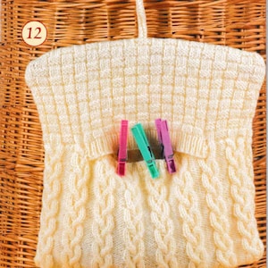May include: Cream-colored knitted garment with a textured pattern and cable knit design. Three colorful clothespins are clipped to the garment. The number 12 is in a red circle in the upper left corner. The garment is hanging on a woven background.