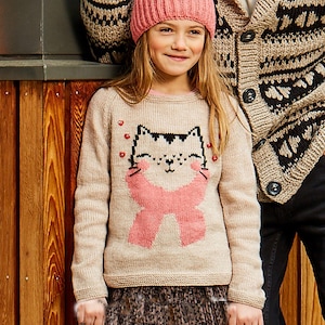 Può includere: Una persona giovane indossa un maglione di maglia beige chiaro con un disegno di faccia di gatto e un fiocco rosa. La persona indossa anche un cappello di maglia rosa e una gonna fantasia. Sullo sfondo, una persona indossa un cardigan fantasia.
