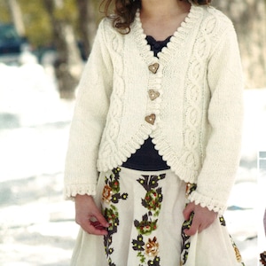 May include: A young person wearing a cream-colored, knitted cardigan with heart-shaped wooden buttons. The cardigan is paired with a floral print skirt and a brown hat. The outfit is suitable for a child.