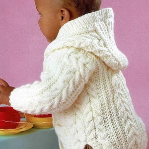 May include: Cream-colored knitted baby hoodie with a cable knit design. The hooded sweater has long sleeves and a split hem. The baby is wearing tan pants. The background is a light pink color.