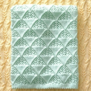 May include: A light blue knitted blanket with a repeating geometric pattern of interlocking arches and triangles. The blanket is folded and lies on a yellow textured surface. The blanket's dimensions are not visible.