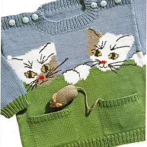 May include: A light blue knitted sweater featuring two white and brown cat faces peeking over a green pocket. A knitted mouse toy is in the pocket. The sweater has light blue buttons on the shoulder.