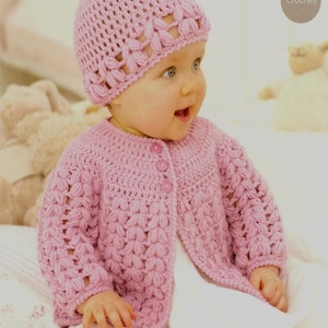 May include: A baby wearing a light pink crochet hat and cardigan. The hat has a floral design around the brim, and the cardigan has three pink buttons. The word "Crochet" is in a circle in the upper right corner.