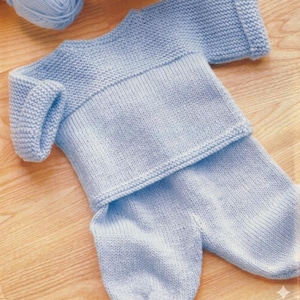 May include: A light blue knitted baby outfit, including a sweater and pants, is displayed on a wooden surface. The outfit is made of soft yarn and features a ribbed collar and cuffs. A ball of matching yarn is visible in the background.