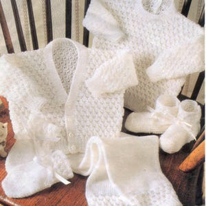 May include: A set of white knitted baby garments, featuring a cardigan, sweater, leggings, and booties with ribbon ties. The items are displayed on a wooden chair, highlighting a textured knit design.