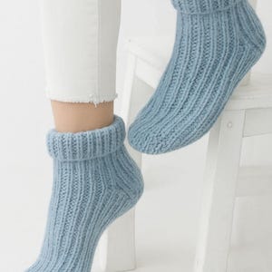 May include: Light blue knitted ankle socks with a ribbed pattern. The socks feature a folded cuff. The image shows the socks being worn with white trousers. The socks are made of a soft, warm material.