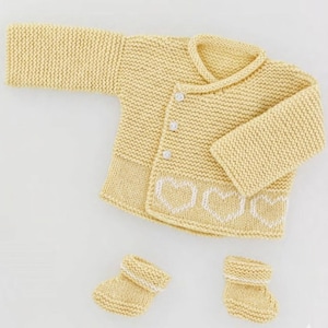 Puede incluir: Conjunto de bebé tejido de color amarillo claro, que incluye una rebeca con detalles de corazones blancos y dos patucos a juego. La rebeca tiene tres botones blancos y un cuello. Los patucos tienen ribetes blancos. El conjunto está hecho de hilo suave.