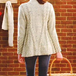 Könnte beinhalten: Ein hellfarbener Zopfstrickpullover mit hohem Kragen und langen Ärmeln. Der Pullover hat ein strukturiertes Design und wird mit dunkelblauen Jeans getragen. Ein geflochtener Korb wird in der Hand gehalten. Der Hintergrund ist eine Backsteinmauer.