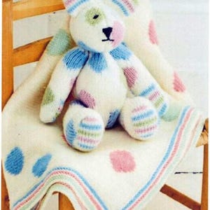May include: A handmade, white knitted teddy bear with blue, pink, and green polka dots and striped accents. The bear is seated on a matching blanket with large polka dots and striped borders. The soft toy is placed on a wooden chair.