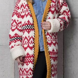 Könnte beinhalten: Lange Strickjacke mit offenem Schnitt, mit einem geometrischen Muster aus Rot und Weiß, mit gelbem Rand und schwarzen Nähten. Die Ärmel haben weiße Manschetten. Die Jacke wird über einem Jeanshemd und einer schwarzen Hose getragen.