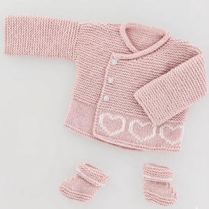 May include: A light pink knitted baby set, including a cardigan with white heart details and two matching booties. The cardigan has three buttons and a collar. The set is made of a soft, textured yarn.