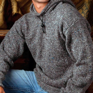 May include: A gray hooded sweater with a speckled pattern. The sweater has a drawstring at the neckline and is paired with blue jeans. The knitwear appears to be thick and warm, suitable for cooler weather.