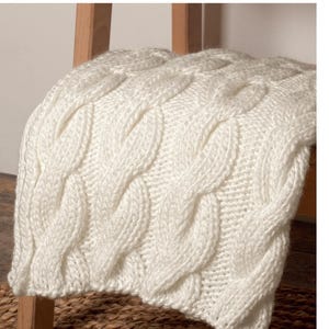 May include: A cream-colored, cable-knit throw blanket draped over a wooden ladder. The blanket features a thick, textured weave with a repeating pattern of raised, intertwined stitches. The neutral color and cozy texture suggest warmth and comfort.
