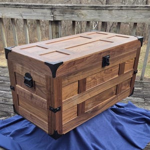 May include: A wooden trunk with a rectangular shape, featuring a warm brown finish. The trunk has black metal corner protectors and a matching latch. The lid is flat with decorative panels. The trunk is sitting on a blue cloth.