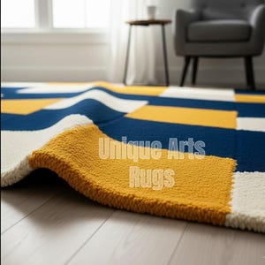 Puede incluir: Primer plano de una alfombra geométrica con un borde doblado. La alfombra presenta un patrón de formas rectangulares azul marino, blancas y amarillas. El texto "Unique Arts Rugs" es visible en la alfombra.