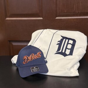 Może przedstawiać: Granatowa czapka bejsbolowa z napisem "Detroit" w pomarańczowym i czarnym skrypcie oraz biała koszulka do baseballu z granatowymi literami i logo "D". Czapka ma metkę "Fanatics".