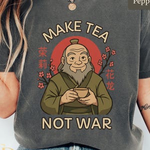 May include: A dark gray t-shirt featuring a cartoon illustration of an elderly man holding a teacup, with the words "MAKE TEA NOT WAR" in white. The design includes a red circle and floral accents, with Chinese characters. The shirt is made of a soft material.