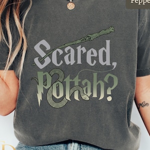May include: A pepper-colored short-sleeve t-shirt featuring the text "Scared, Pottah?" in a stylized font. The design includes a green wand graphic positioned above the text and a green snake graphic integrated into the lettering of the word Pottah.