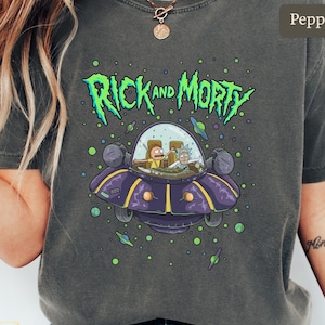 May include: A gray t-shirt featuring the text "Rick and Morty" in green, with a purple spaceship graphic. The spaceship contains cartoon characters. The shirt is a comfortable, casual wear item.