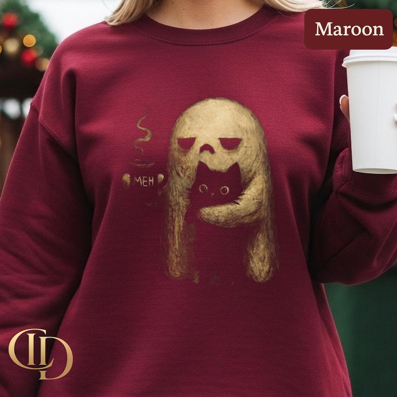 May include: Maroon sweatshirt featuring a gold illustration of a skull-like figure embracing a black cat. A coffee cup with steam and the word "MEH" are also depicted. The sweatshirt is a solid maroon color.