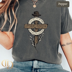 May include: Pepper-coloured t-shirt with a graphic design featuring the text "Arthur Morgan" on a cross-shaped emblem. The design includes the phrases "Blessed are those who hunger" and "and thirst for righteousness."