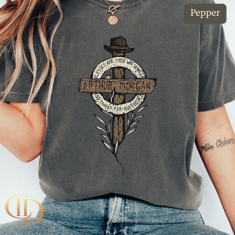 May include: Pepper-coloured t-shirt with a graphic design featuring the text "Arthur Morgan" on a cross-shaped emblem. The design includes the phrases "Blessed are those who hunger" and "and thirst for righteousness."
