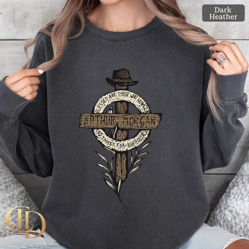 May include: Dark heather grey sweatshirt with a graphic of a cross and a hat. The cross has the name "Arthur Morgan" on it, with the text "Blessed are those who hunger and thirst for righteousness" around it.