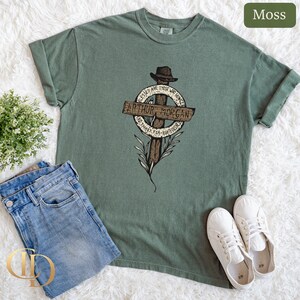 May include: A moss green t-shirt with a graphic featuring a cross, hat, and the text "Arthur Morgan". The shirt is styled with blue jeans and white trainers. The image presents casual clothing.