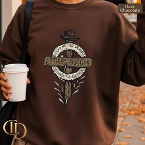 May include: Dark chocolate-coloured sweatshirt with a graphic design. The design features a cross with a hat, the name "Arthur Morgan", and the text "Blessed are those who hunger and thirst for righteousness."