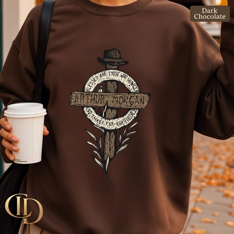 May include: Dark chocolate-coloured sweatshirt with a graphic design. The design features a cross with a hat, the name "Arthur Morgan", and the text "Blessed are those who hunger and thirst for righteousness."
