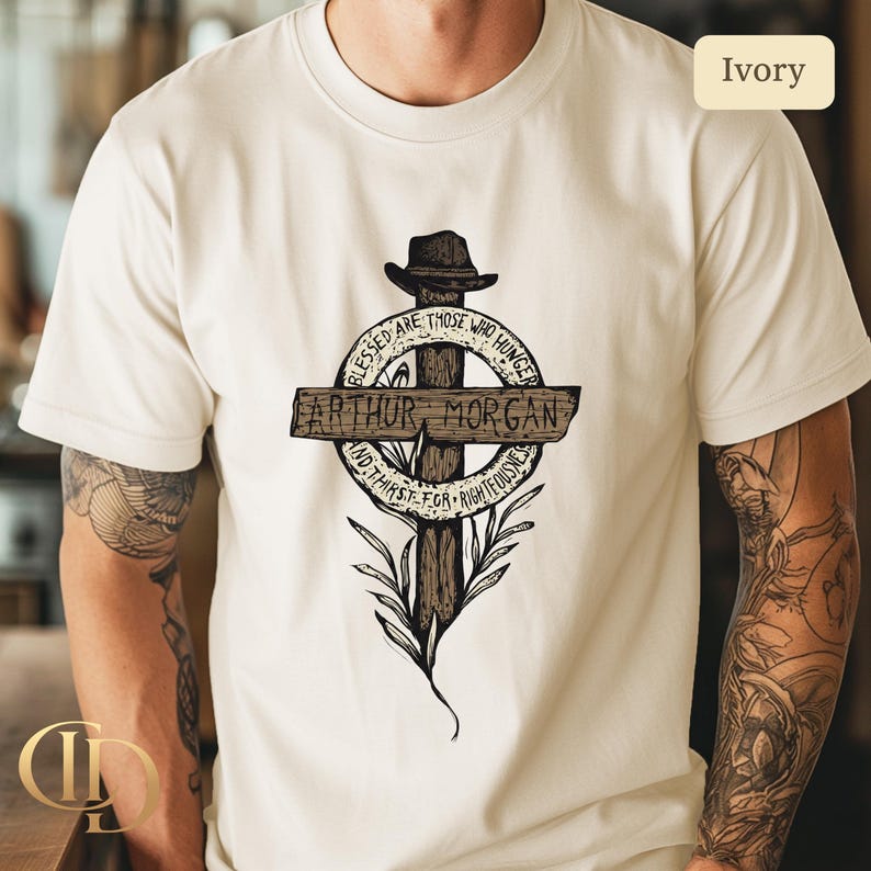 May include: Ivory t-shirt with a graphic design featuring a cross, a hat, and the text "ARTHUR MORGAN". The design includes the phrases "Blessed are those who hunger" and "And thirst for righteousness".