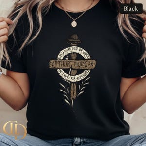 May include: Black t-shirt with a graphic design featuring the text "ARTHUR MORGAN" on a wooden cross. The design includes the quote "BLESSED ARE THOSE WHO HUNGER AND THIRST FOR RIGHTEOUSNESS". The shirt has a crew neck and short sleeves.