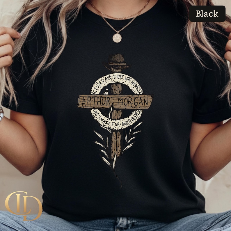 May include: Black t-shirt with a graphic design featuring the text "ARTHUR MORGAN" on a wooden cross. The design includes the quote "BLESSED ARE THOSE WHO HUNGER AND THIRST FOR RIGHTEOUSNESS". The shirt has a crew neck and short sleeves.