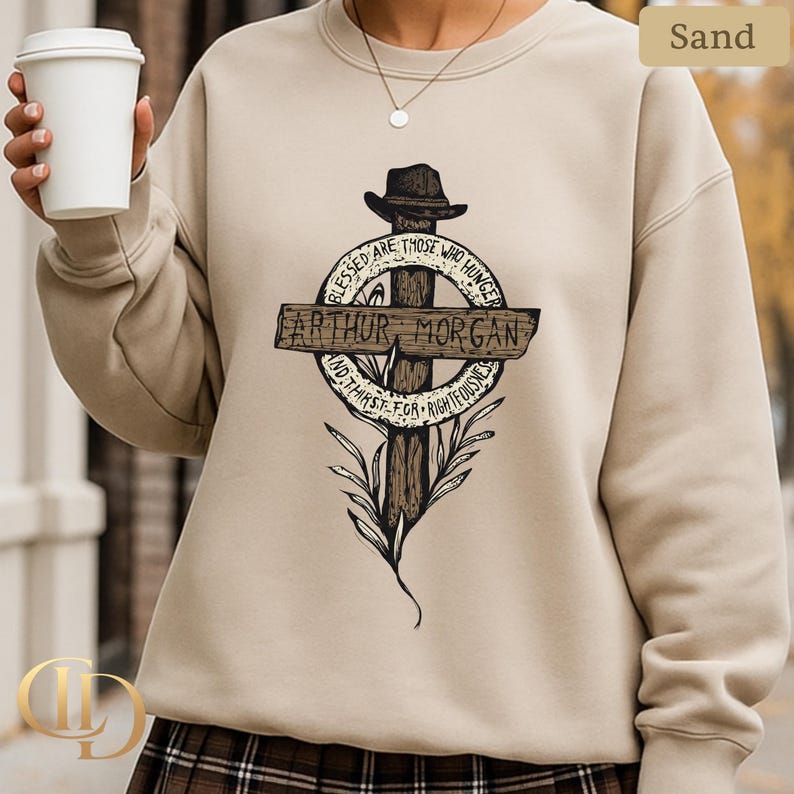 May include: Sand-coloured sweatshirt with a graphic of a cross, a hat, and a circular design with text. The text reads "Blessed are those who hunger and thirst for righteousness" and includes the name "Arthur Morgan."