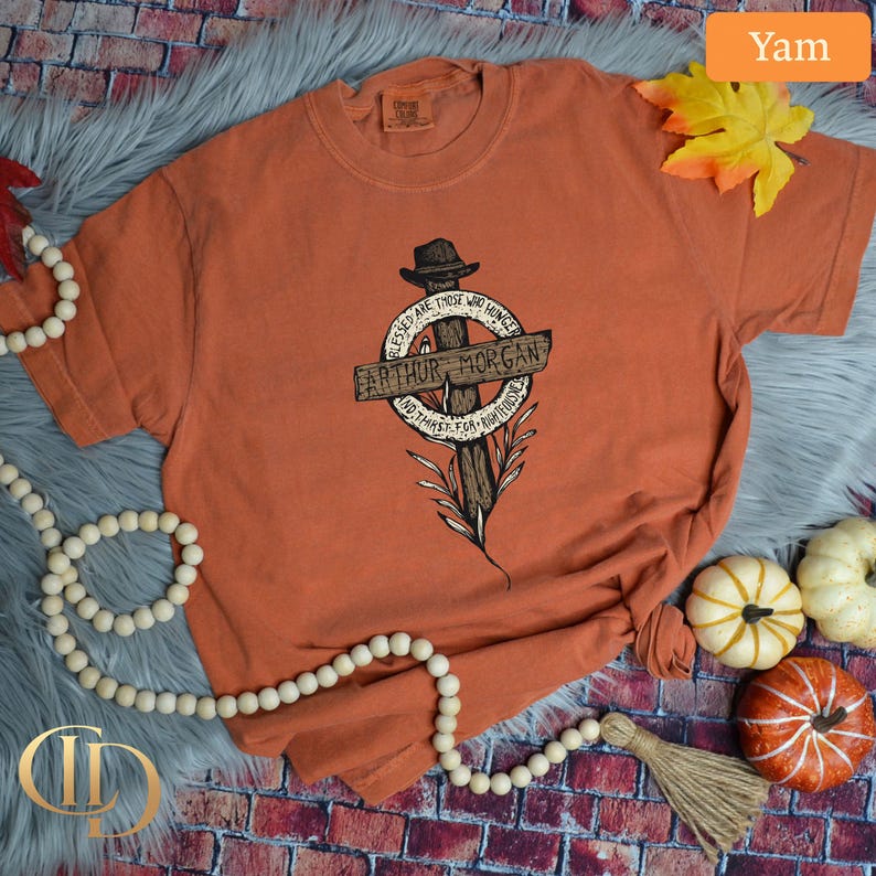 May include: A yam-coloured t-shirt featuring a graphic of a cross with a hat and the text "Arthur Morgan". The shirt also has the text "Blessed are those who hunger and thirst for righteousness".