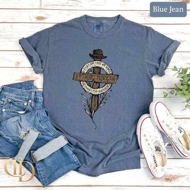 May include: Blue jean coloured t-shirt featuring a graphic of a cross, a hat, and the words "Arthur Morgan". The shirt is shown with blue jeans and white trainers.