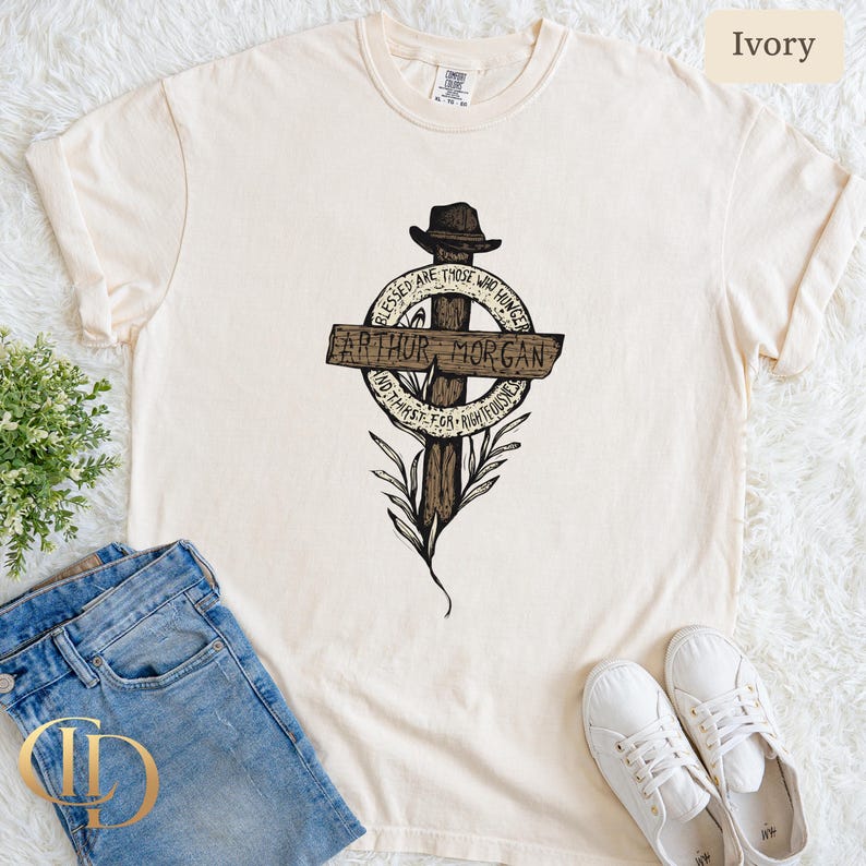 May include: Cream-coloured t-shirt with a graphic of a cross, a hat, the text "ARTHUR MORGAN", and the quote "Blessed are those who hunger and thirst for righteousness". The shirt is styled with blue jeans and white trainers.