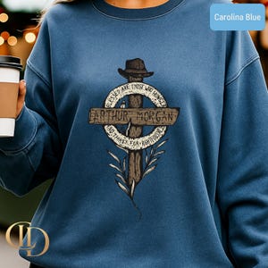 May include: Carolina blue sweatshirt with a cross graphic, a hat, and the text "ARTHUR MORGAN". The design includes the words "Blessed are those who hunger and thirst for righteousness". The graphic is in brown and white.