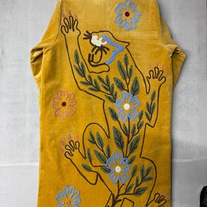May include: Mustard yellow velvet coat featuring an embroidered panther design with blue, green, and pink floral accents. The coat has long sleeves and a relaxed fit, perfect for layering. The panther is outlined in brown with pink claws.