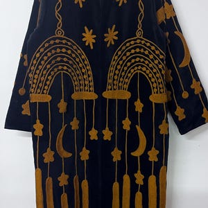 Pode incluir: Um casaco preto de manga comprida com bordados em amarelo mostarda. O design apresenta motivos de arco-íris arqueados, estrelas pendentes, luas crescentes e formas retangulares verticais. A peça tem gola alta e fecho de correr frontal.