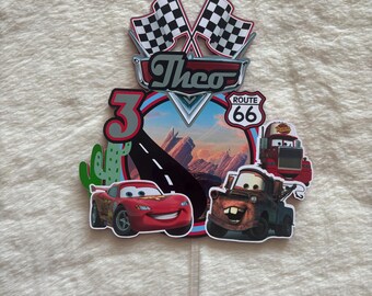 Può includere: Un colorato cake topper con i personaggi del film Cars, tra cui Saetta McQueen e Cricchetto. Il topper presenta il nome "Theo" e il numero "3", insieme a bandiere a scacchi e un cartello della Route 66.