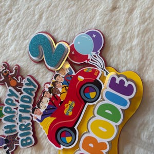 Pu&ograve; includere: Topper colorato per torta di compleanno con un'auto rossa dei Wiggles, un numero 2 blu glitterato e palloncini. Il topper include anche le parole "Happy Birthday" e "RODDIE".