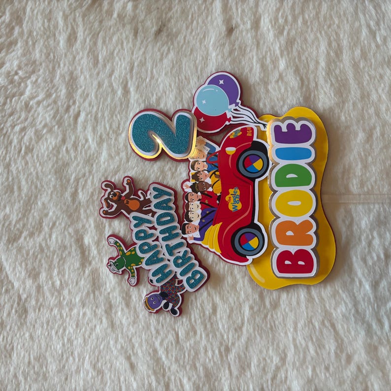 Pu&ograve; includere: Topper colorato per torta di compleanno con il numero 2 glitterato, un'auto rossa con i personaggi dei Wiggles e il nome "Brodie". Include un messaggio "Buon Compleanno" e palloncini.