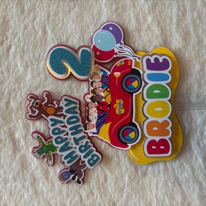 Pu&ograve; includere: Decorazione colorata per torta di compleanno con un'auto rossa con i personaggi dei Wiggles, un numero 2 blu glitterato, palloncini e il nome "Brodie". Include anche un cane e una rana. Sono incluse anche le parole "Happy Birthday".
