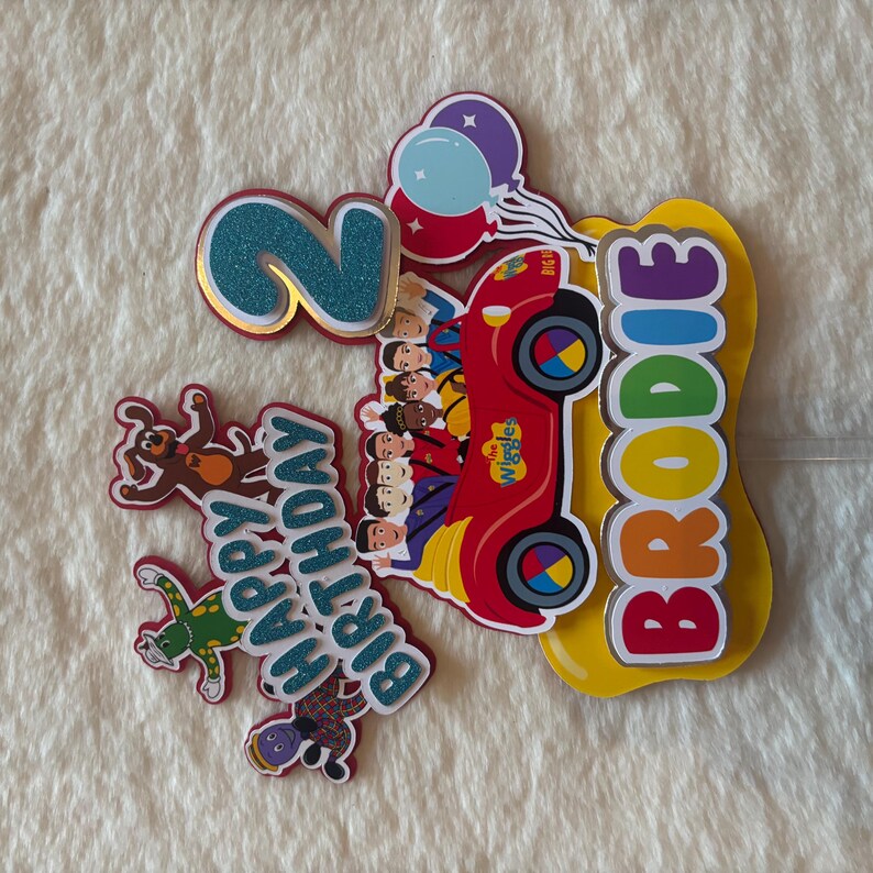 Pu&ograve; includere: Decorazione colorata per torta di compleanno con un'auto rossa con i personaggi dei Wiggles, un numero 2 blu glitterato, palloncini e il nome "Brodie". Include anche un cane e una rana. Sono incluse anche le parole "Happy Birthday".