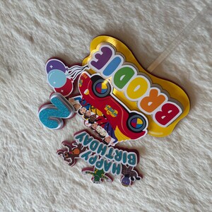 Pu&ograve; includere: Una collezione di decorazioni colorate per torte di compleanno. Include un'auto rossa con la scritta "The Wiggles", il nome "Brodie", il numero "2", palloncini e la frase "Happy Birthday".